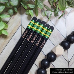 May include: Five black pencils, each inscribed with "Kolten", are displayed. The pencils feature green and yellow bands near the eraser. The erasers are grey. The background includes greenery and a black beaded accessory.