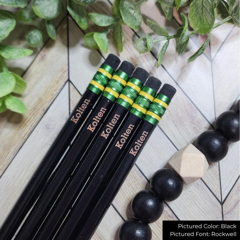 May include: Five black pencils, each inscribed with "Kolten", are displayed. The pencils feature green and yellow bands near the eraser. The erasers are grey. The background includes greenery and a black beaded accessory.