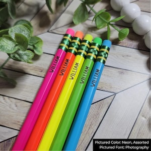 May include: A selection of neon-coloured pencils, each inscribed with the name "WILLIAM". The pencils feature pink, orange, yellow, green, and blue barrels, complemented by matching erasers. They are displayed on a wooden surface.