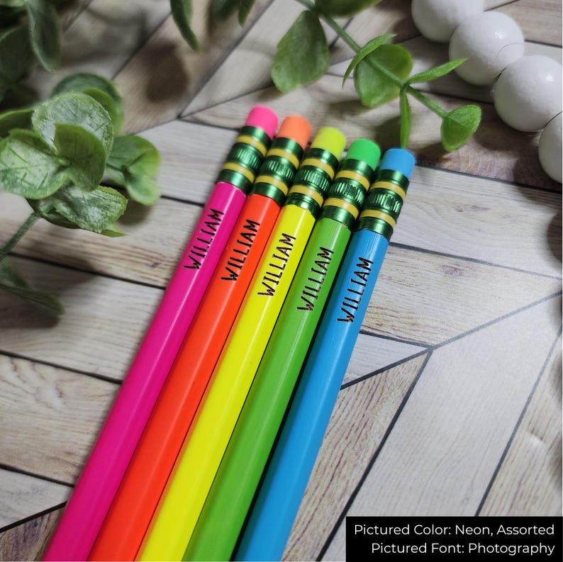 May include: A selection of neon-coloured pencils, each inscribed with the name "WILLIAM". The pencils feature pink, orange, yellow, green, and blue barrels, complemented by matching erasers. They are displayed on a wooden surface.
