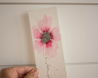 Pode incluir: Uma flor rosa em aquarela com um centro preto e branco e as palavras "Eu te amo" escritas em tinta preta.