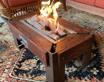 May include: A wooden fire pit table with a metal firebox and a flame burning inside. The table has a dark brown stain and is sitting on a colorful rug.