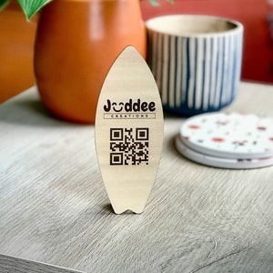 以下が含まれることがあります： 「Jodddee Creations」というテキストと白黒のQRコードが書かれた木製のサーフボード型の看板です。