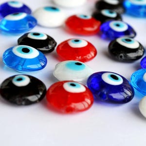 May include: A collection of glass evil eye beads in various colors, including blue, red, black, and white. Each bead features a central blue eye design with a black pupil. The beads are scattered on a white surface.