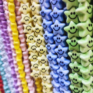 May include: Rows of colorful star-shaped beads with smiling faces. The beads are in shades of yellow, blue, and green, with other colors visible in the background. The beads are strung together, creating a textured and playful pattern.