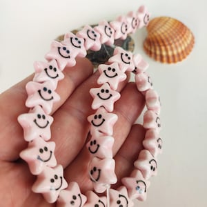 May include: A strand of pink star-shaped beads with black smiling faces. The beads are held in a hand, with a seashell in the background. The beads are approximately 0.5 inches in diameter.