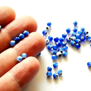 May include: A collection of small, round, blue glass beads with a white and black eye design. Some beads are held in a hand, while others are scattered on a white surface. These beads are often used in jewelry making.