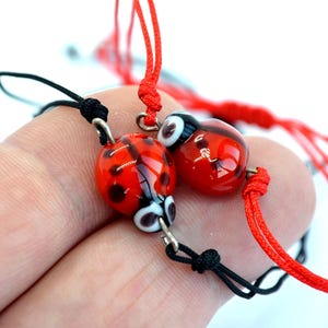 May include: Two ladybug-shaped glass bead bracelets. The ladybugs are red with black spots and white and black eyes. The bracelets have red and black cords.