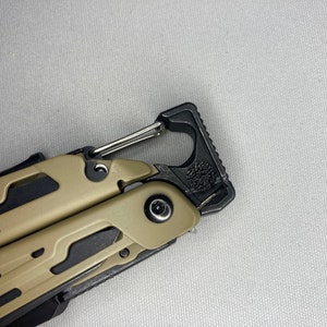 Puede incluir: Una multiherramienta de color marrón claro y negro. La herramienta tiene un mosquetón y la palabra "SIGNAL" está impresa en el lateral.