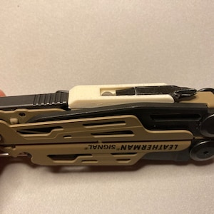 Peut inclure: Un multi-outil Leatherman Signal de couleur beige et noir avec un accessoire en plastique blanc sur le dessus. Le multi-outil comprend une variété d'outils, notamment un couteau, une scie, une lime et un tournevis.
