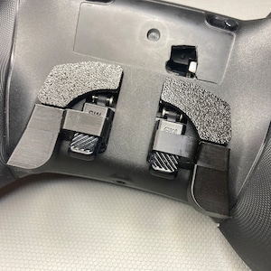 May include: A black gaming controller with two black paddle attachments on the back. The paddles are labelled "CM1" and "CM2".