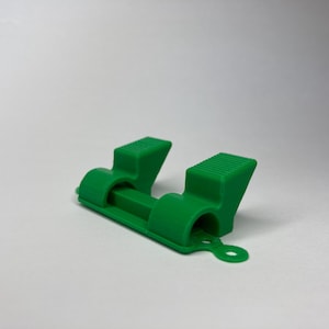 Puede incluir: Una herramienta de plástico verde con dos puntas, diseñada para sujetar o asegurar objetos.