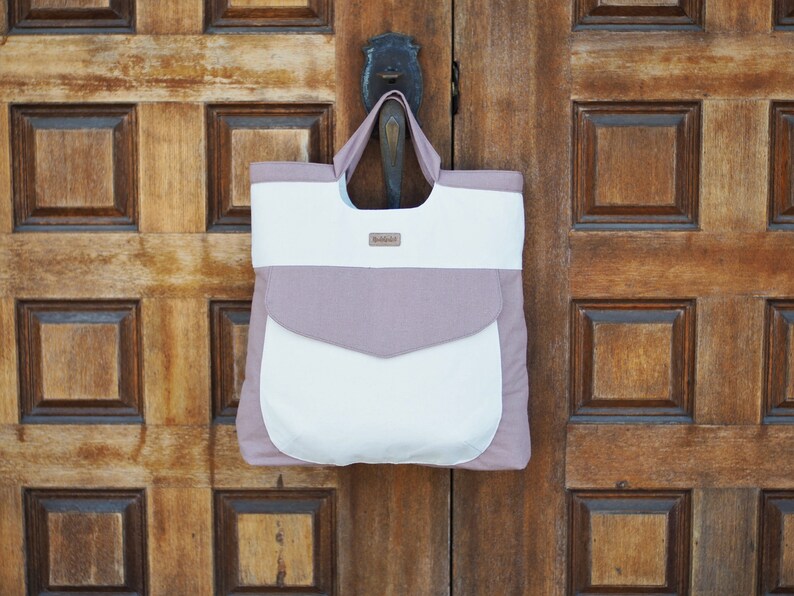 May include: A white and light pink tote bag with a pocket on the front. The bag has a brown leather tag with the word "Wildfolk" on it.