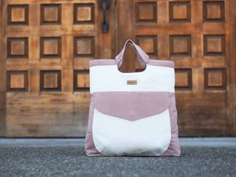 May include: A pink and white tote bag with a large pocket on the front. The bag has two handles and a small brown label with the word "Babble" on it.