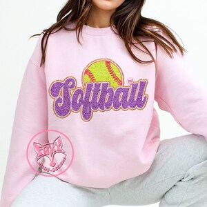 May include: A pink sweatshirt with a purple glitter design that says "Softball" with a yellow softball graphic.