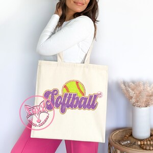 May include: A white canvas tote bag with a purple and gold glitter design that says "Softball" with a yellow softball graphic.
