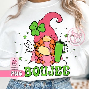May include: A pink and green gnome with a shamrock on its hat, holding a green tumbler and a bottle. The gnome is sitting on a green and white background with the word "BOUJEE" written in green letters.