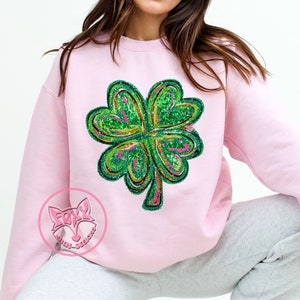 Peut inclure: Un sweat-shirt rose avec un motif de tr&egrave;fle &agrave; quatre feuilles vert en paillettes. Le tr&egrave;fle est bord&eacute; d'or et de rose.
