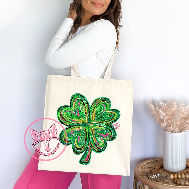 Peut inclure: Un sac fourre-tout en toile blanche avec un motif de tr&egrave;fle &agrave; quatre feuilles vert. Le tr&egrave;fle est fait de paillettes vertes et a un contour rose. Le sac a un logo rose et blanc qui dit "Foxy-Brain Designs".