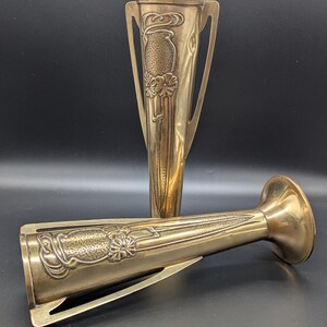 May include: Two antique brass vases with intricate floral designs. The vases are both tall and slender, with a flared base and a narrow neck. The designs are embossed and feature a raised floral motif.