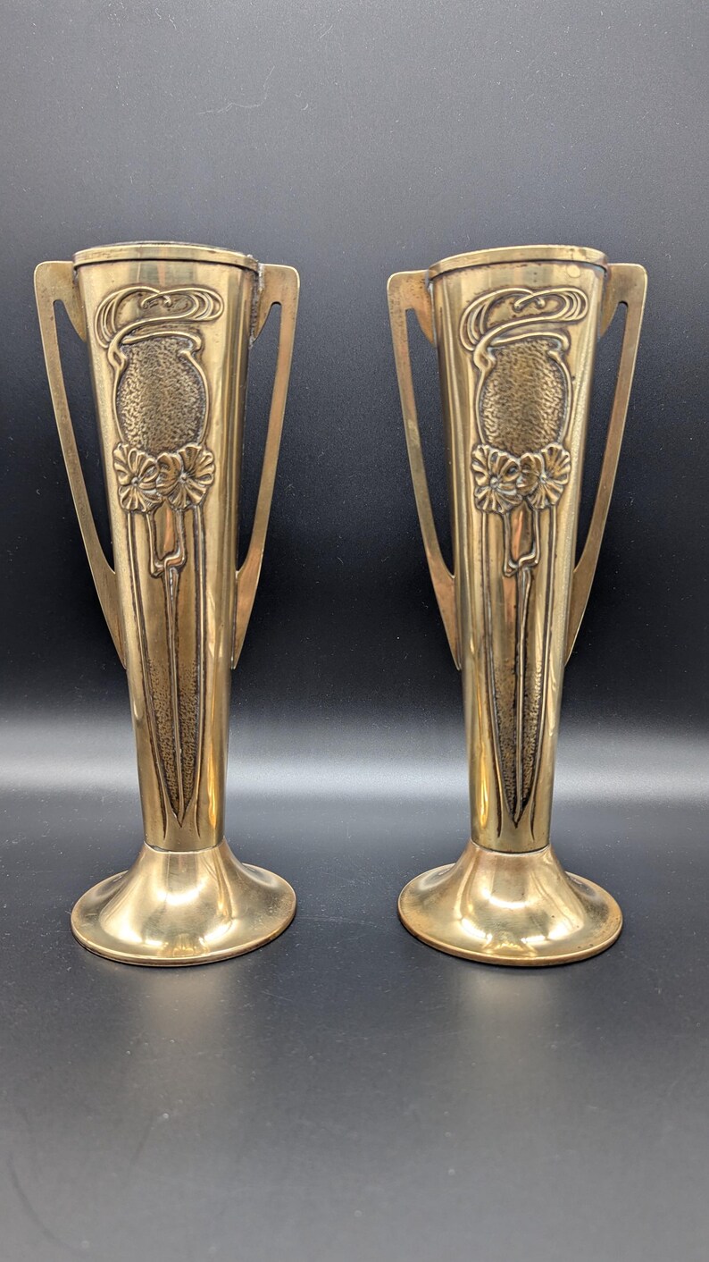 May include: A pair of antique brass vases with ornate floral designs and two handles. The vases are tall and slender with a flared base.