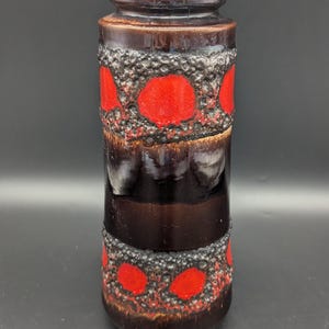 Puede incluir: Un jarrón cilíndrico de cerámica con un esmalte marrón oscuro. El jarrón presenta bandas de material texturizado gris oscuro con acentos circulares rojos. El jarrón mide aproximadamente 25 cm de alto.