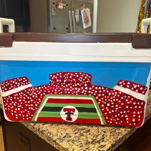 May include: A rectangular cooler with a brown top, white handles, and a blue body. The cooler is painted with a stadium design in red and white, with a green and black emblem featuring a red "T" in a circle. The cooler is designed for outdoor events.