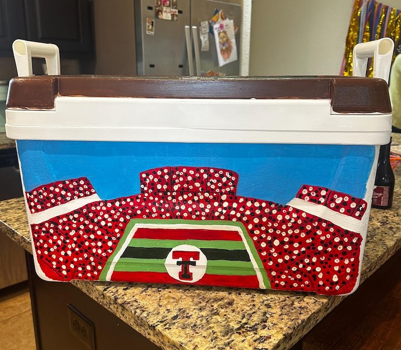 May include: A rectangular cooler with a brown top, white handles, and a blue body. The cooler is painted with a stadium design in red and white, with a green and black emblem featuring a red "T" in a circle. The cooler is designed for outdoor events.