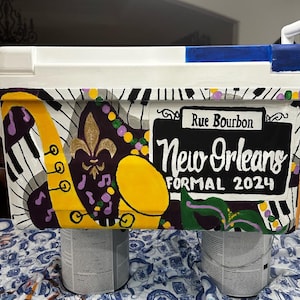 May include: A white cooler with a hand-painted New Orleans theme. The design includes a yellow saxophone, piano keys, a fleur-de-lis, and the text "Rue Bourbon" and "New Orleans Formal 2024". The cooler is supported by two paint cans.