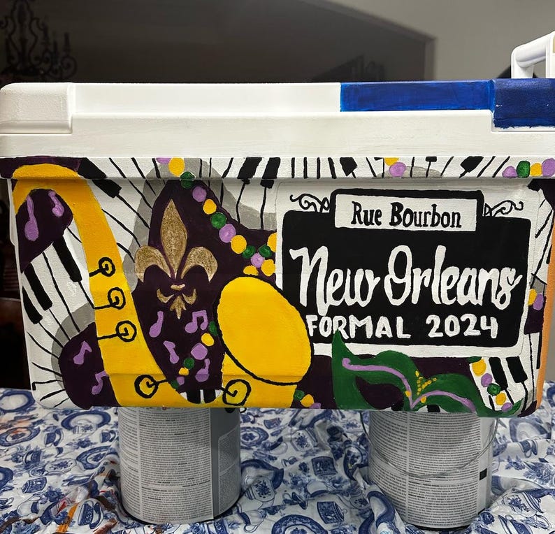 May include: A white cooler with a hand-painted New Orleans theme. The design includes a yellow saxophone, piano keys, a fleur-de-lis, and the text "Rue Bourbon" and "New Orleans Formal 2024". The cooler is supported by two paint cans.