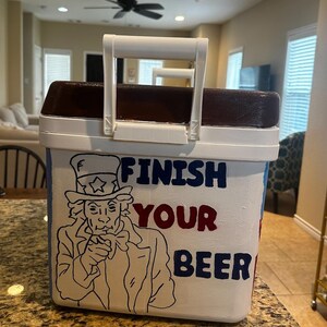 May include: White cooler with a brown top and a white handle. The cooler features a drawing of Uncle Sam and the words "FINISH YOUR BEER" in blue and red lettering.