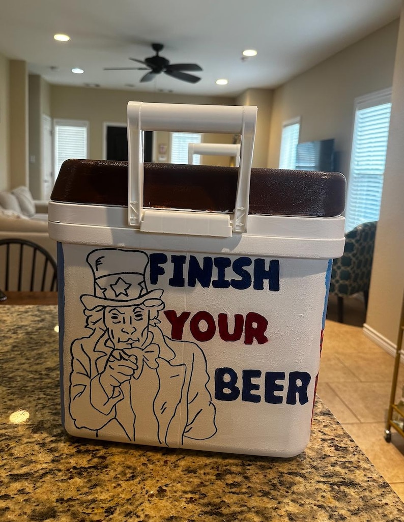 May include: White cooler with a brown top and a white handle. The cooler features a drawing of Uncle Sam and the words "FINISH YOUR BEER" in blue and red lettering.