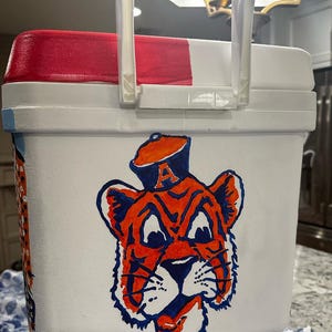 May include: White cooler with a red and white lid and a clear plastic handle. The cooler features a hand-painted orange and blue tiger mascot design on the front. The tiger is wearing a hat with the letter "A".