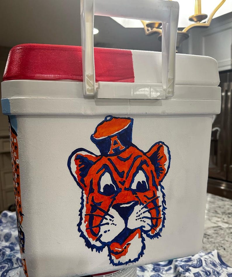 May include: White cooler with a red and white lid and a clear plastic handle. The cooler features a hand-painted orange and blue tiger mascot design on the front. The tiger is wearing a hat with the letter "A".