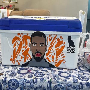 May include: A white and blue cooler with a portrait of a man and orange abstract shapes. The cooler has white handles and the text "Mikey Burton" on the lid. The OVO owl logo is also visible.