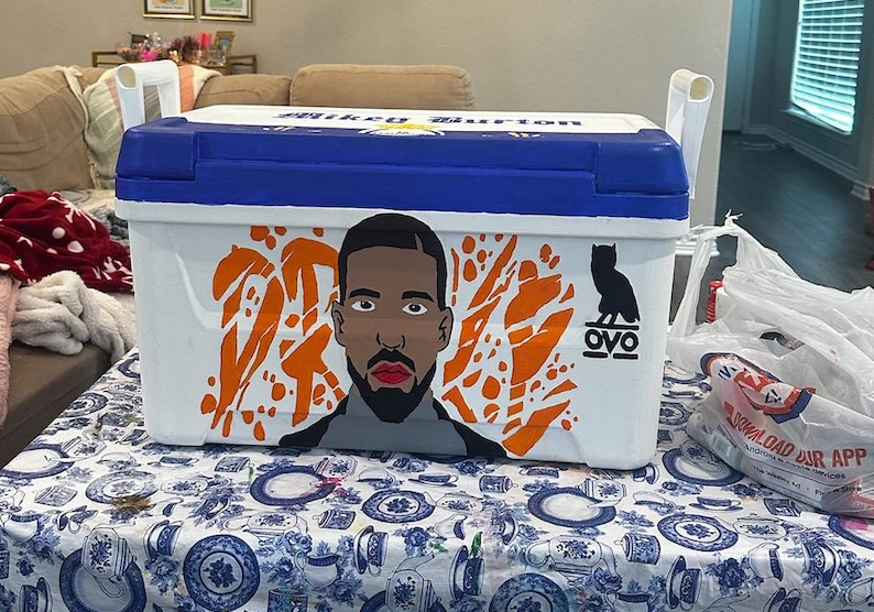 May include: A white and blue cooler with a portrait of a man and orange abstract shapes. The cooler has white handles and the text "Mikey Burton" on the lid. The OVO owl logo is also visible.