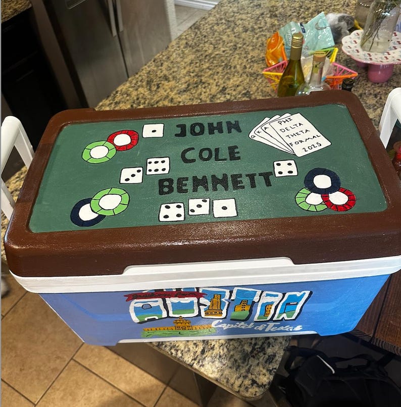 May include: A blue and brown cooler with a painted poker table design on the lid. The lid features dice, poker chips, and the name "JOHN COLE BENNETT". The cooler also has a "Capital of Texas" design.