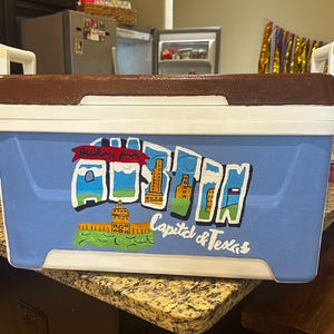 May include: A rectangular cooler with a blue body, white trim, and a brown lid. The cooler features a hand-painted design with the words "Greetings from" and "Capital & Texas" along with illustrations of landmarks.
