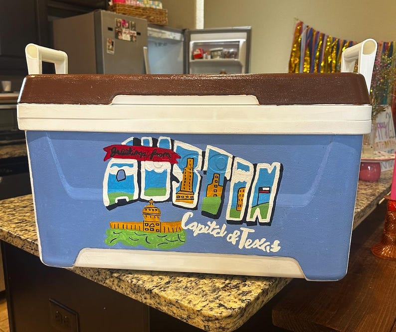 May include: A rectangular cooler with a blue body, white trim, and a brown lid. The cooler features a hand-painted design with the words "Greetings from" and "Capital & Texas" along with illustrations of landmarks.
