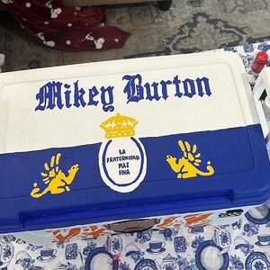 May include: A white and blue cooler with the name "Mikey Burton" in ornate lettering. The cooler features a blue lower section with yellow emblem designs and the text "LA FRATERNIDAD MAS FINA" in a circular emblem.
