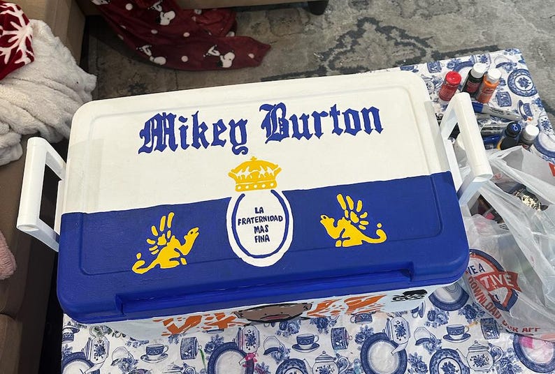 May include: A white and blue cooler with the name "Mikey Burton" in ornate lettering. The cooler features a blue lower section with yellow emblem designs and the text "LA FRATERNIDAD MAS FINA" in a circular emblem.