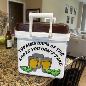 May include: White and brown cooler with a white handle. The cooler has the text "YOU MISS 100% OF THE SHOTS YOU DON'T TAKE" with a graphic of two shot glasses and lime wedges.