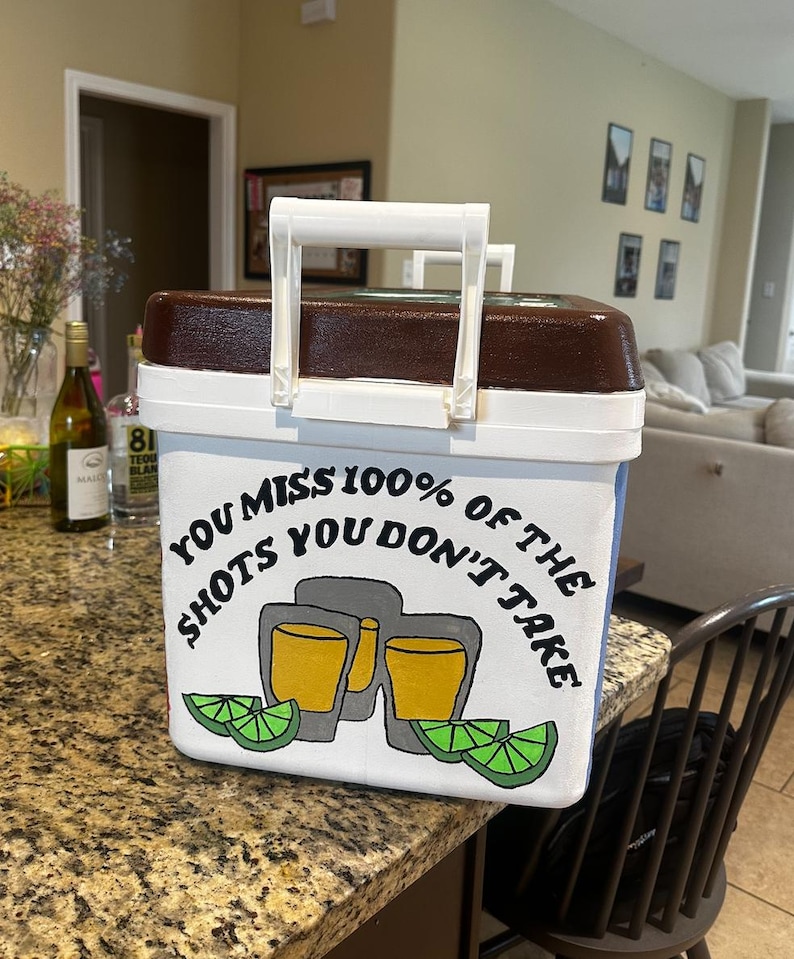 May include: White and brown cooler with a white handle. The cooler has the text "YOU MISS 100% OF THE SHOTS YOU DON'T TAKE" with a graphic of two shot glasses and lime wedges.