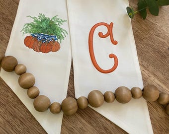 May include: Two off-white fabric pieces with embroidered fall designs. One features pumpkins and a fern in a blue and white pot. The other has an orange letter "C". A wooden bead garland is in the foreground.
