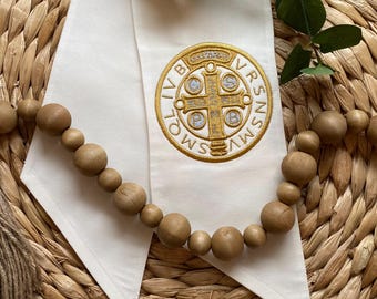 May include: White fabric with a gold embroidered St. Benedict medal. The medal has the words "VRSNSMVS" and "IVBVS" around the edge. The fabric is on a woven straw surface.