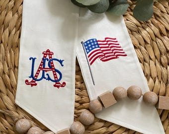 May include: Two off-white fabric pennants featuring embroidered designs. One pennant displays a red and blue monogram of the letters US, while the other features a small, waving American flag on a pole. The fabric is smooth and light-colored.