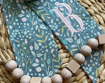 May include: A pair of green fabric ribbon with a floral pattern and the letter "B" embroidered on one of the ribbons. The ribbons are decorated with a string of wooden beads and blocks.