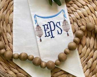 May include: White fabric with blue embroidered initials "EPS" and decorative elements. The design includes a blue banner, two hanging lantern-like figures, and a brown tassel. A wooden bead garland is in the foreground.