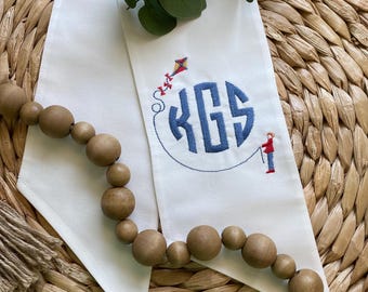 May include: White cloth napkin with a blue monogram and a kite design. The monogram features the letters KGS in a circular design. A small embroidered figure is holding the kite string. A wooden bead garland is in the foreground.
