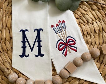 May include: Two off-white fabric ribbons featuring embroidery. One ribbon displays a large navy blue letter M in a serif font. The other ribbon features an embroidered design of three firework rockets tied together with a red, white, and blue bow.